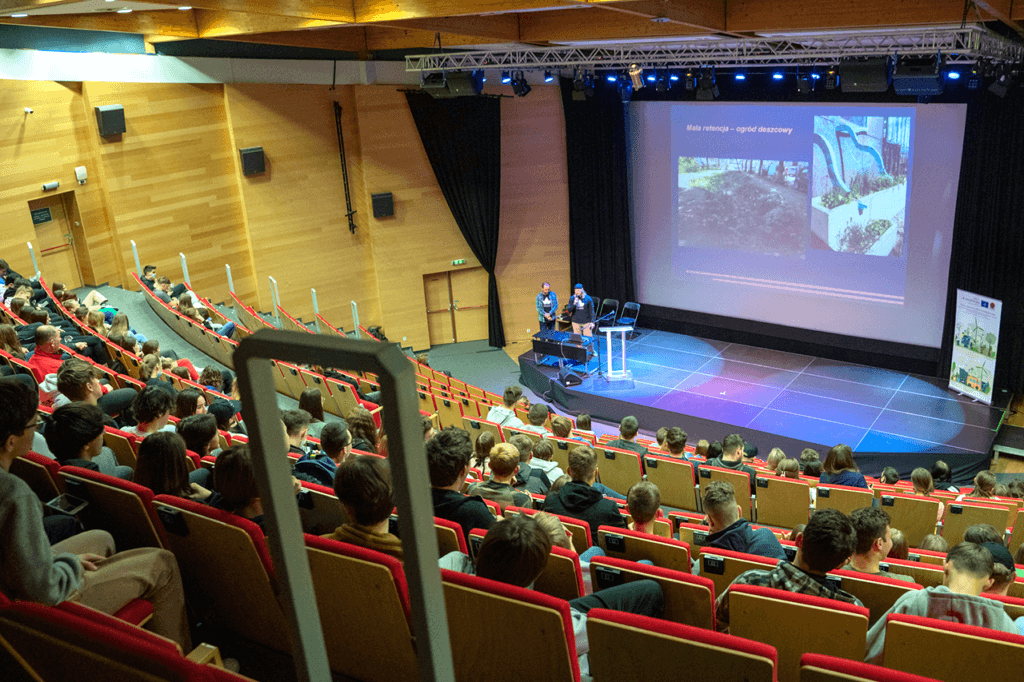 Zdjęcie przedstawia salę wykładową. Na krzesłach siedzą uczestnicy uważnie słuchający eksperta AerisFuturo prowadzącego wykład o edukacji ekologicznej.