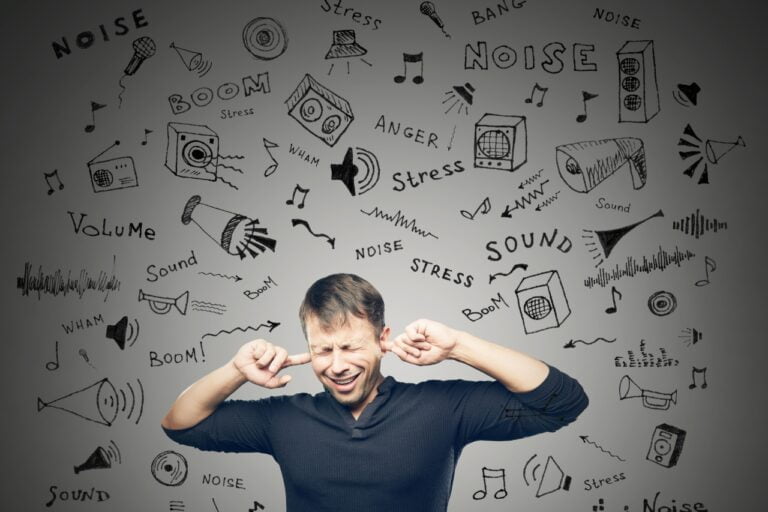Na zdjęciu widzimy mężczyznę z zamkniętymi oczami, ubranego w ciemną koszulę, który zakrywa uszy palcami. Wokół niego znajdują się różne odręczne ilustracje i słowa związane z hałasem oraz dźwiękiem. Można dostrzec głośniki, mikrofony, nuty muzyczne, fale dźwiękowe, a także wyrazy takie jak „NOISE”, „Volume”, „Stress”, „Sound”, „Boom”, „Anger”, „WHAM” i „BANG”. Obraz wizualnie przedstawia koncepcję przytłoczenia przez hałas i dźwięki, podkreślając stres oraz dyskomfort, jaki mogą wywoływać.