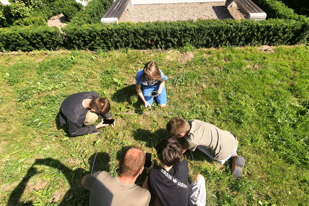 Na zdjęciu widoczne dzieci kucające na trawniku. Razem z prowadzącym warsztaty botaniczne, dzieci identyfikują rośliny znajdujące się wewnątrz niewielkiego kwadratu wyznaczonego na trawniku przy pomocy sznurka. Jest to tak zwane zdjęcie fitosocjologiczne.
