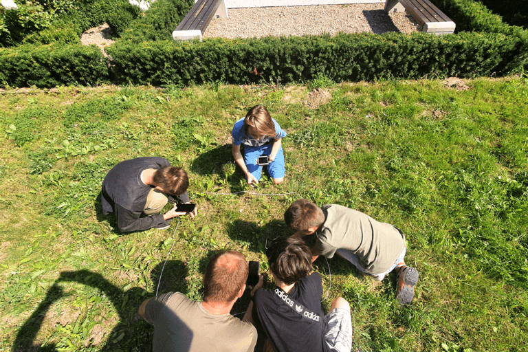 Na zdjęciu widoczne dzieci kucające na trawniku. Razem z prowadzącym warsztaty botaniczne, dzieci identyfikują rośliny znajdujące się wewnątrz niewielkiego kwadratu wyznaczonego na trawniku przy pomocy sznurka. Jest to tak zwane zdjęcie fitosocjologiczne.