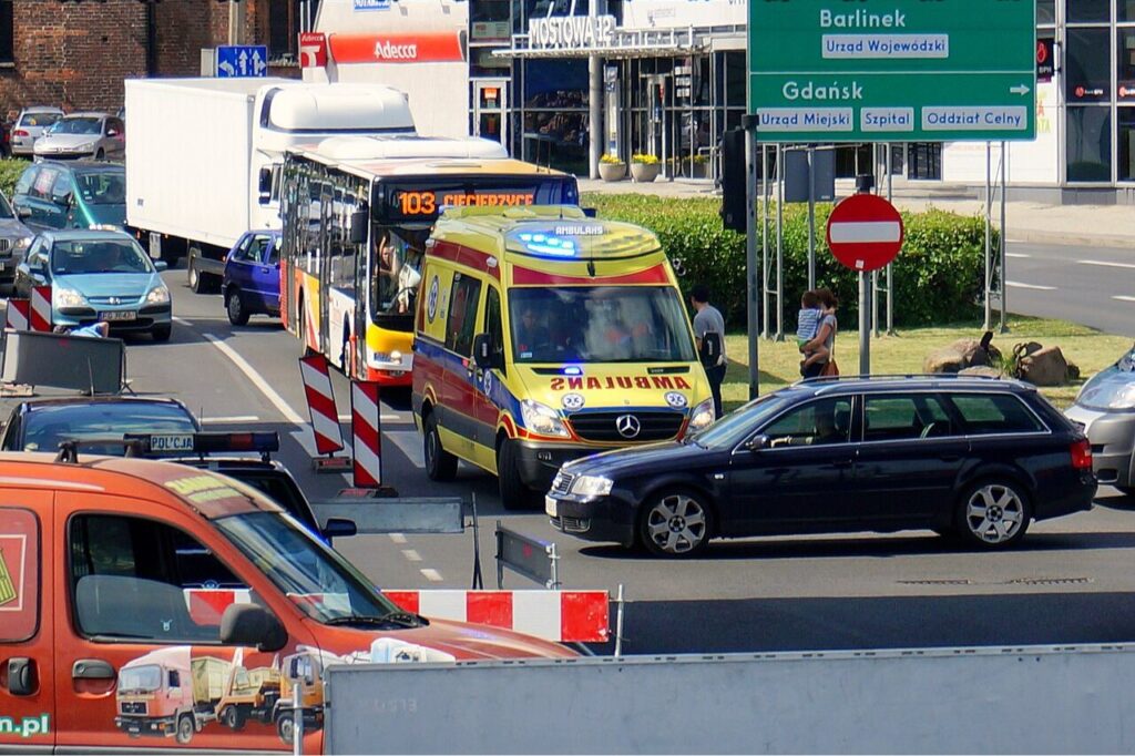 Na zdjęciu widoczna zakorkowana ulica. Możemy dostrzec pogotowie na sygnałach, ludzi, samochody. 