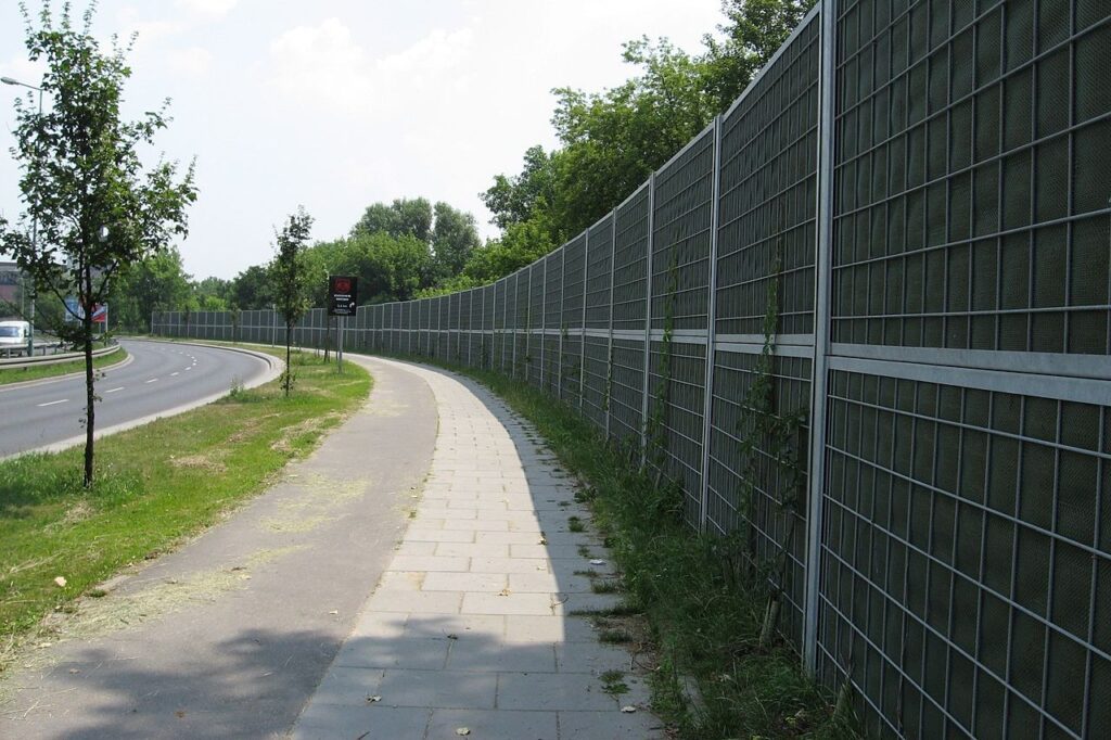 na zdjęciu widoczna ulica, po jej prawej stronie znajduje się chodnik i panek akustyczny, który ma na celu wyciszyć od hałasu pobliskie tereny.